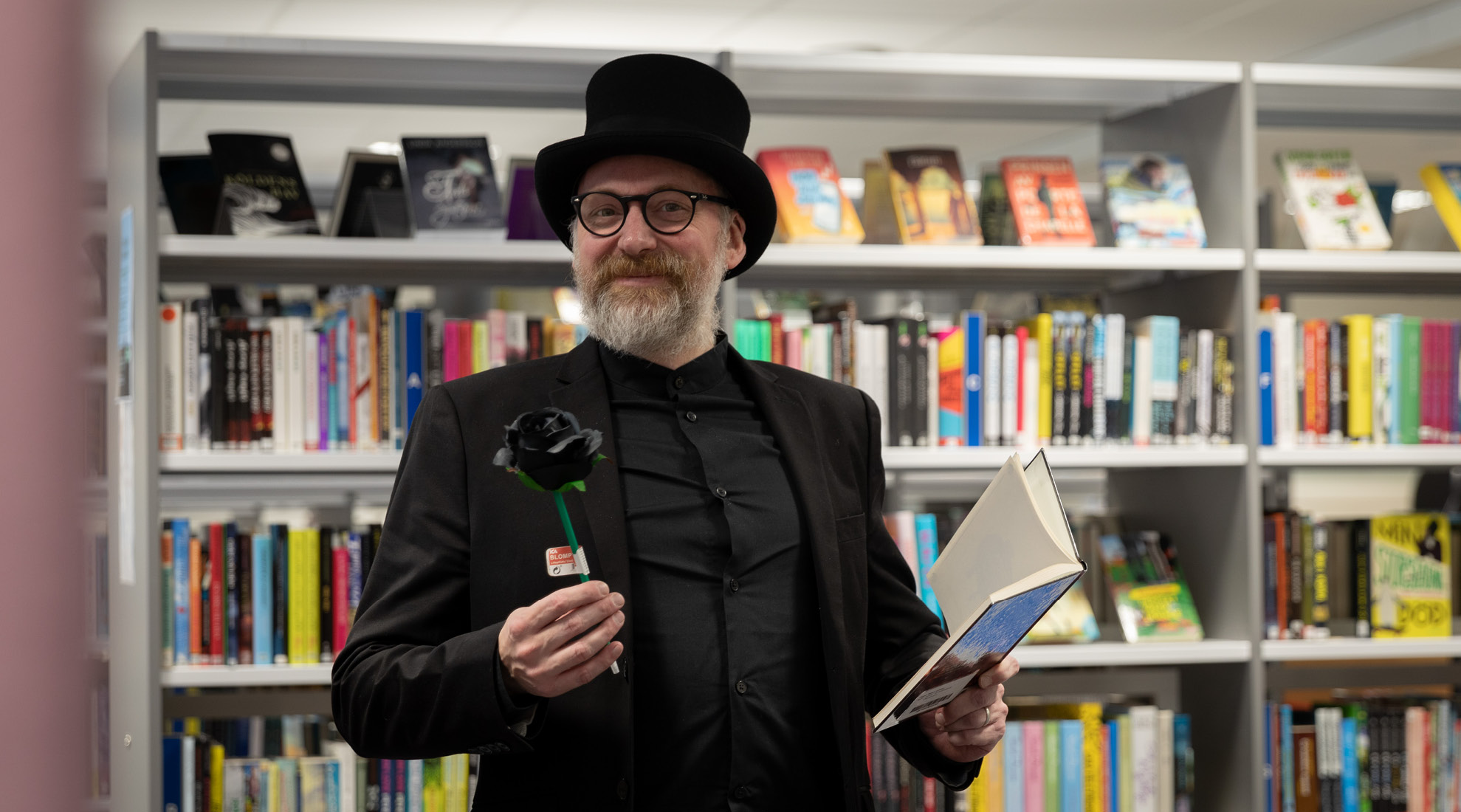 Erling Persson står bland böcker. Han har en svart hatt och håller i en ros och en bok.