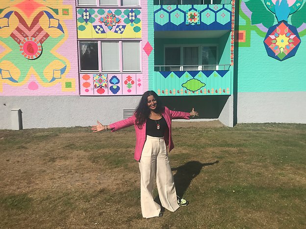 Konstnären Saadia Hussain breder ut armarna åt sidorna framför husväggen hon har målat. Foto: Anna-Karin Sjökvist