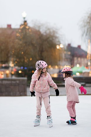 Två barn åker skridskor på isbanan på torget. Foto: De har rosa jackor och rosa hjälmar. Hanna Maxstad.